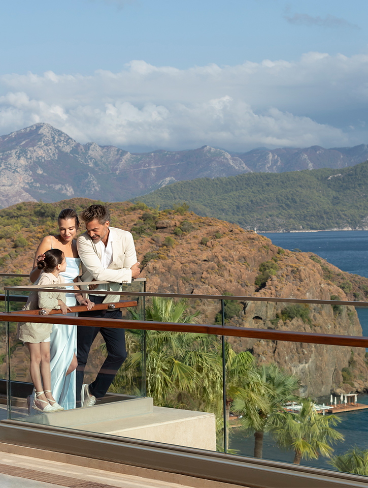  A small family relax on a balcony at the D Maris Bay resort. Mother and Father gaze at their daughter, while behind them is the beautiful ocean and mountain landscape.