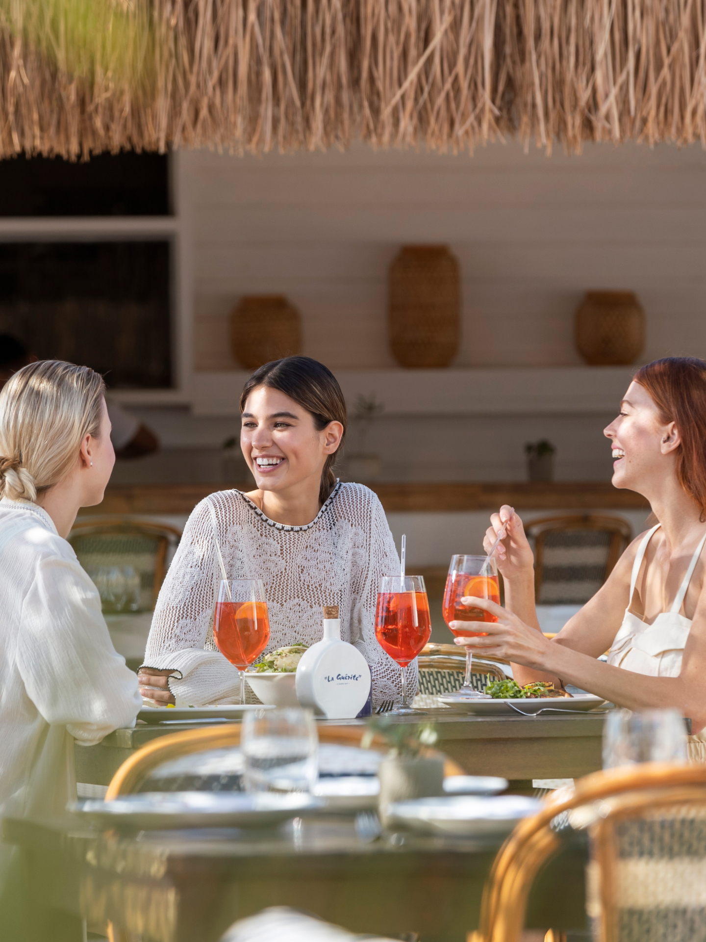 Three women enjoy cocktails and food at La Guerite restaurant at the D Maris Bay resort. Two of the women stand up and dance while the third woman cheers them on. 
