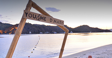 A wooden sign reading ‘You and me D Maris’ in front of a sunset over the sea and mountain view.